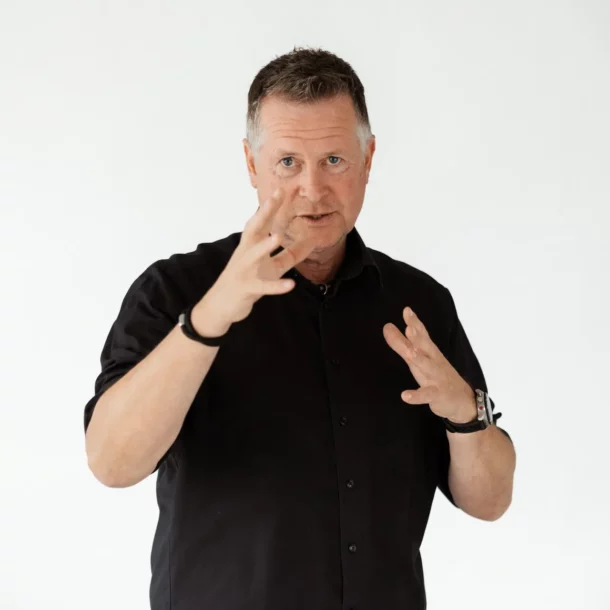 Im schimmernden weißen Raum steht Rhetoriktrainer Ike Fast in einfangender Pose, seinen Blick der Kamera zugewandt.