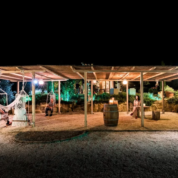 Auf dem Bild sieht man eine Holzüberdachung unter der mehrere Personen gemütlich sitzen, in einem Garten, bei Nacht.