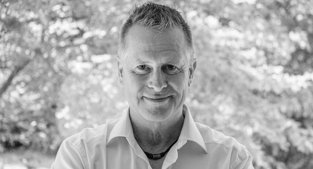 Freundlicher Rhetoriktrainer Ike Fast lächelt in Nahaufnahme zur Kamera, umgeben von Natur, in Graustufen. "Blog" Header.